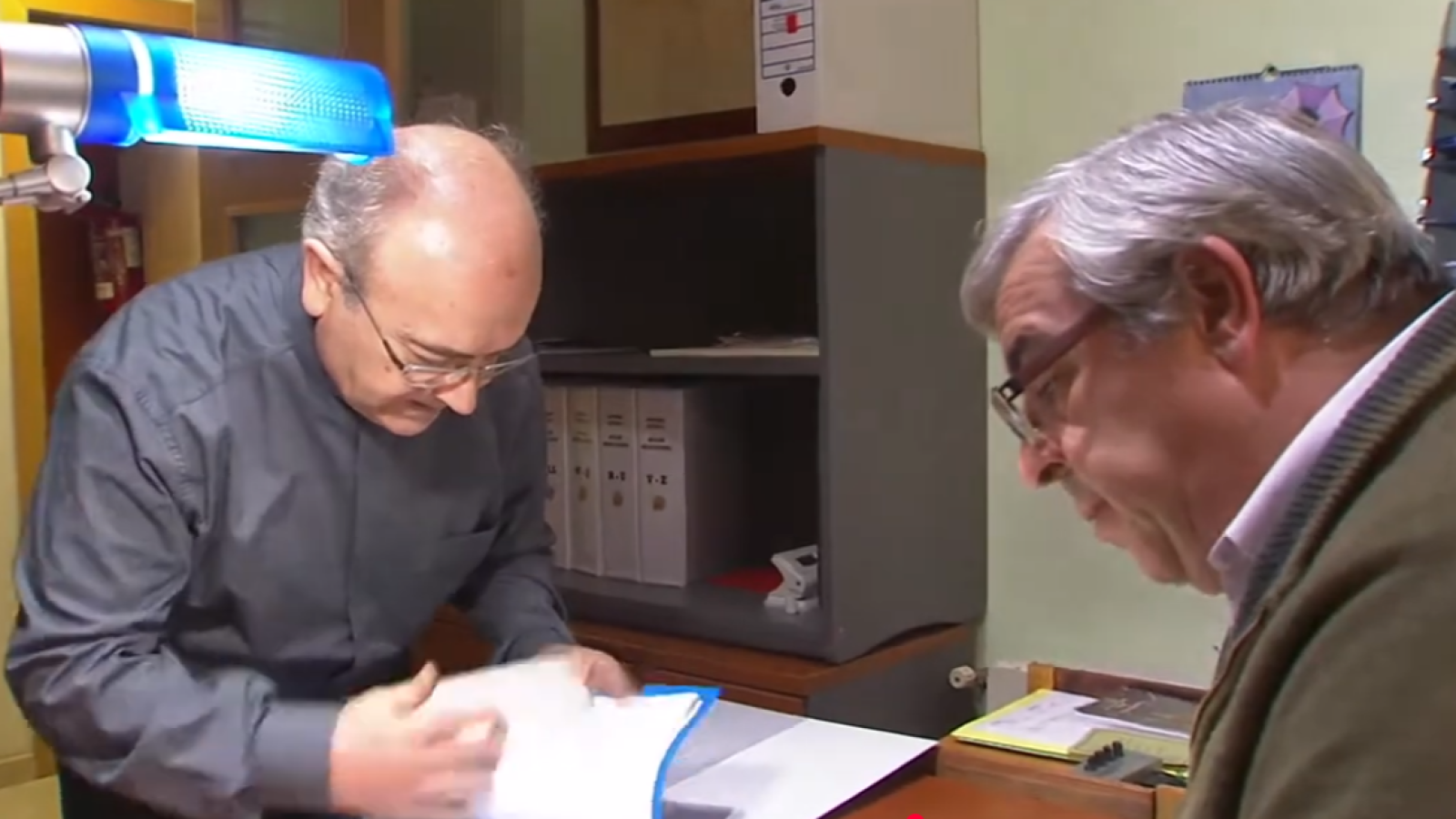 José María Carod y Jesús Bel, ambos con gafas, revisan documentos en un despacho. Una lámpara azul ilumina a Carod, quien manipula papeles en una carpeta azul.