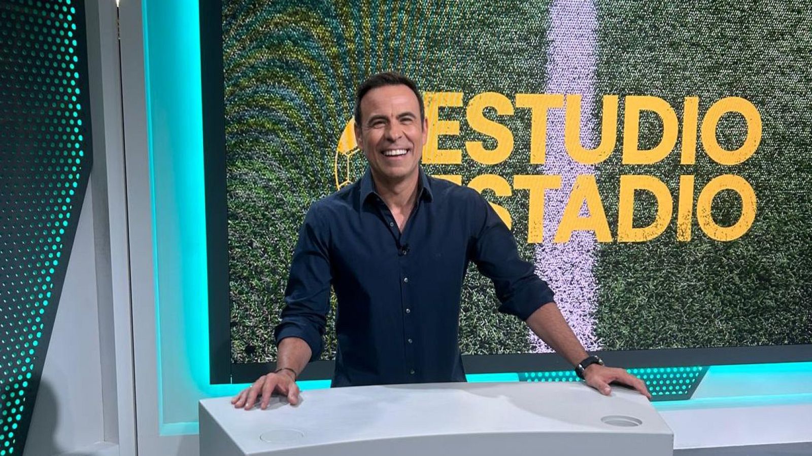 Felipe del Campo, presentador de 'Estudio Estadio', sonríe en un estudio con fondo de campo de fútbol y el nombre del programa en amarillo.