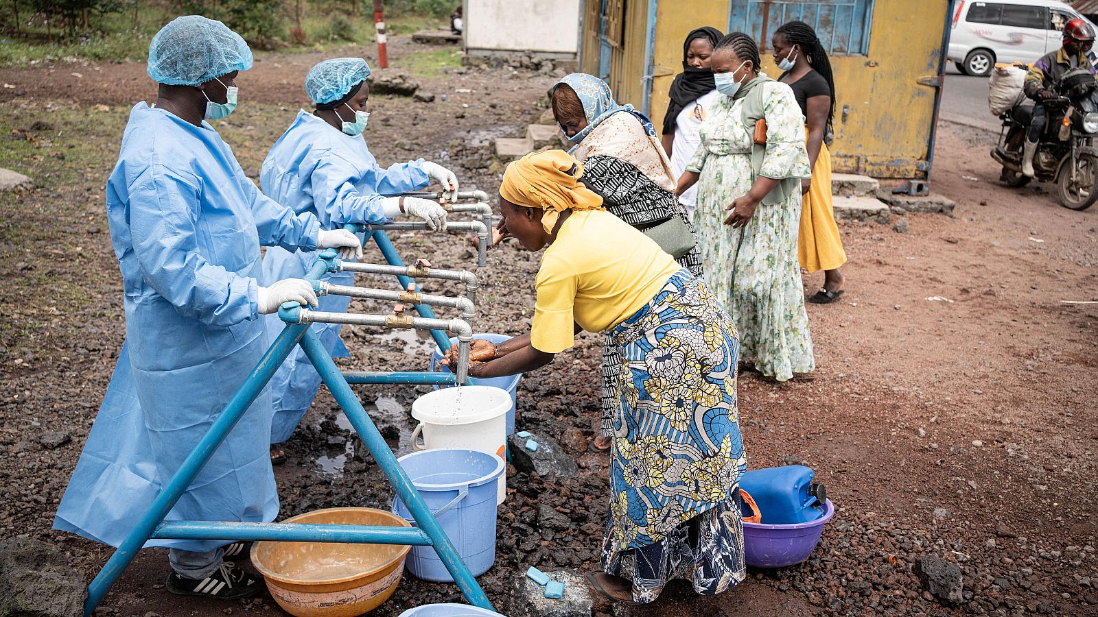 Puesto de control de higiene en R.D. del Congo: habitantes locales se lavan las manos con jabón bajo grifos, supervisados por personal con trajes protectores, como medida preventiva contra el ébola.