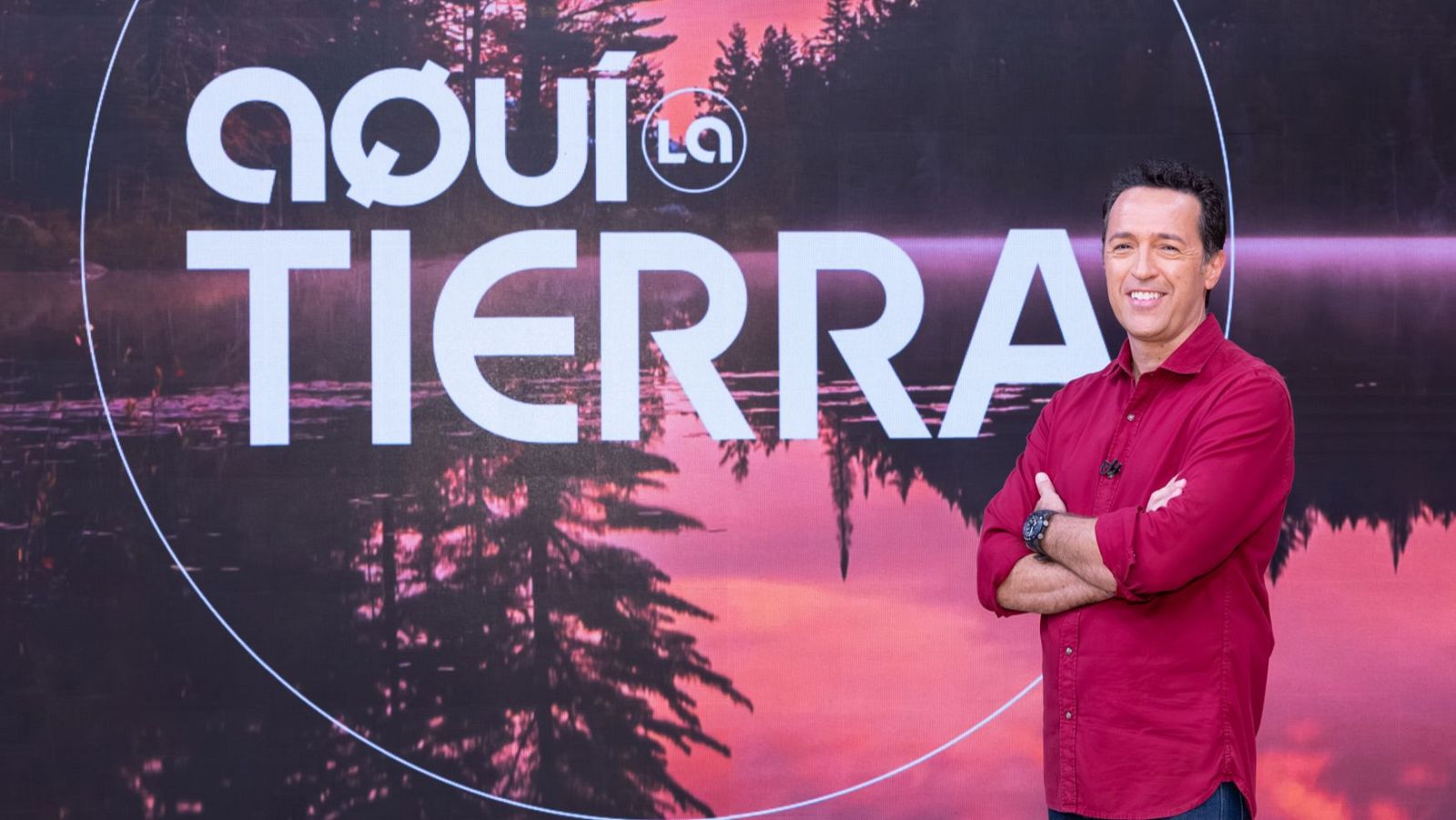 Jacob Petrus, presentador de "Aquí la Tierra", sonríe a cámara con los brazos cruzados, luciendo una camisa granate. El fondo muestra un paisaje natural con el título del programa y el logo de "La 1".