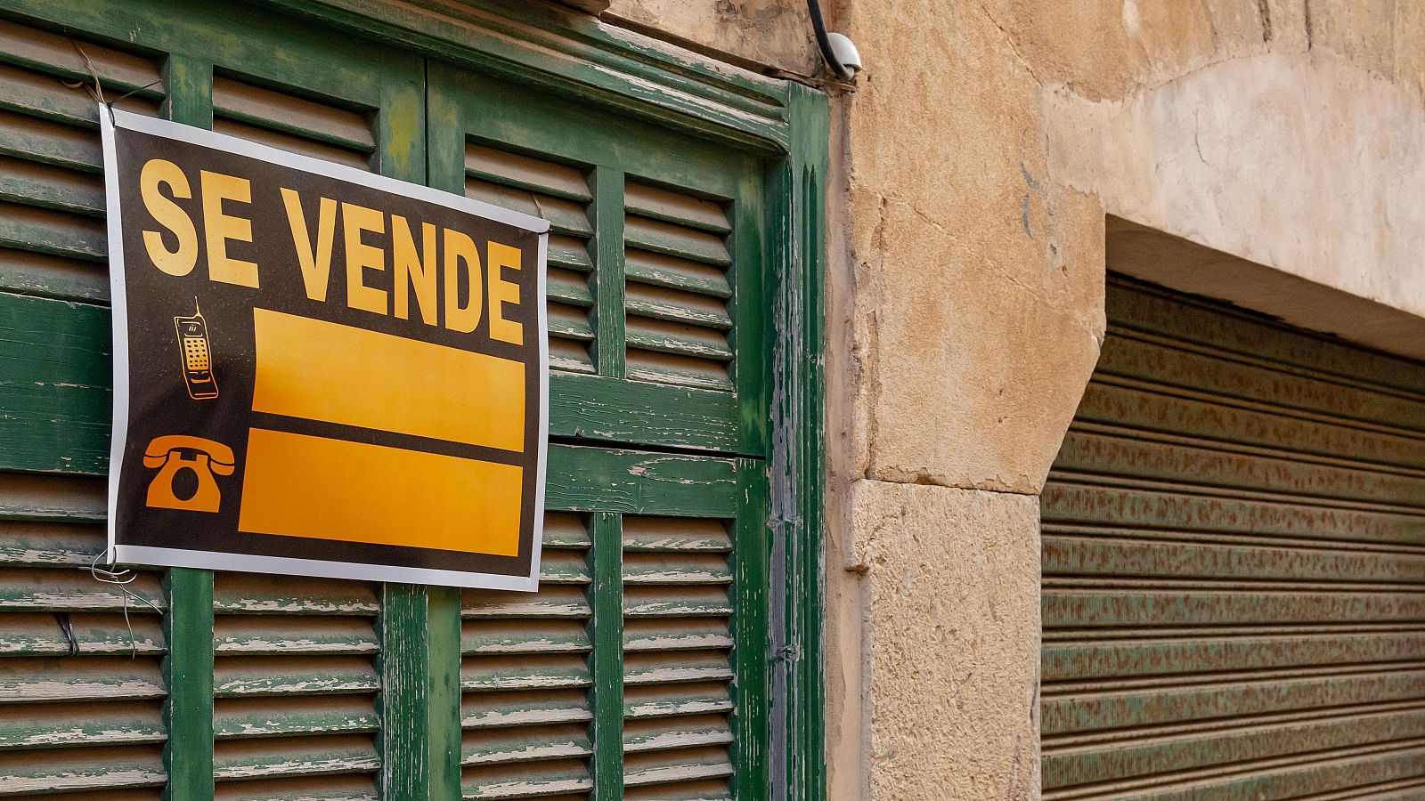 Cartel amarillo de "SE VENDE" con iconos de teléfono sobre persianas verdes desgastadas en la fachada de un edificio antiguo.