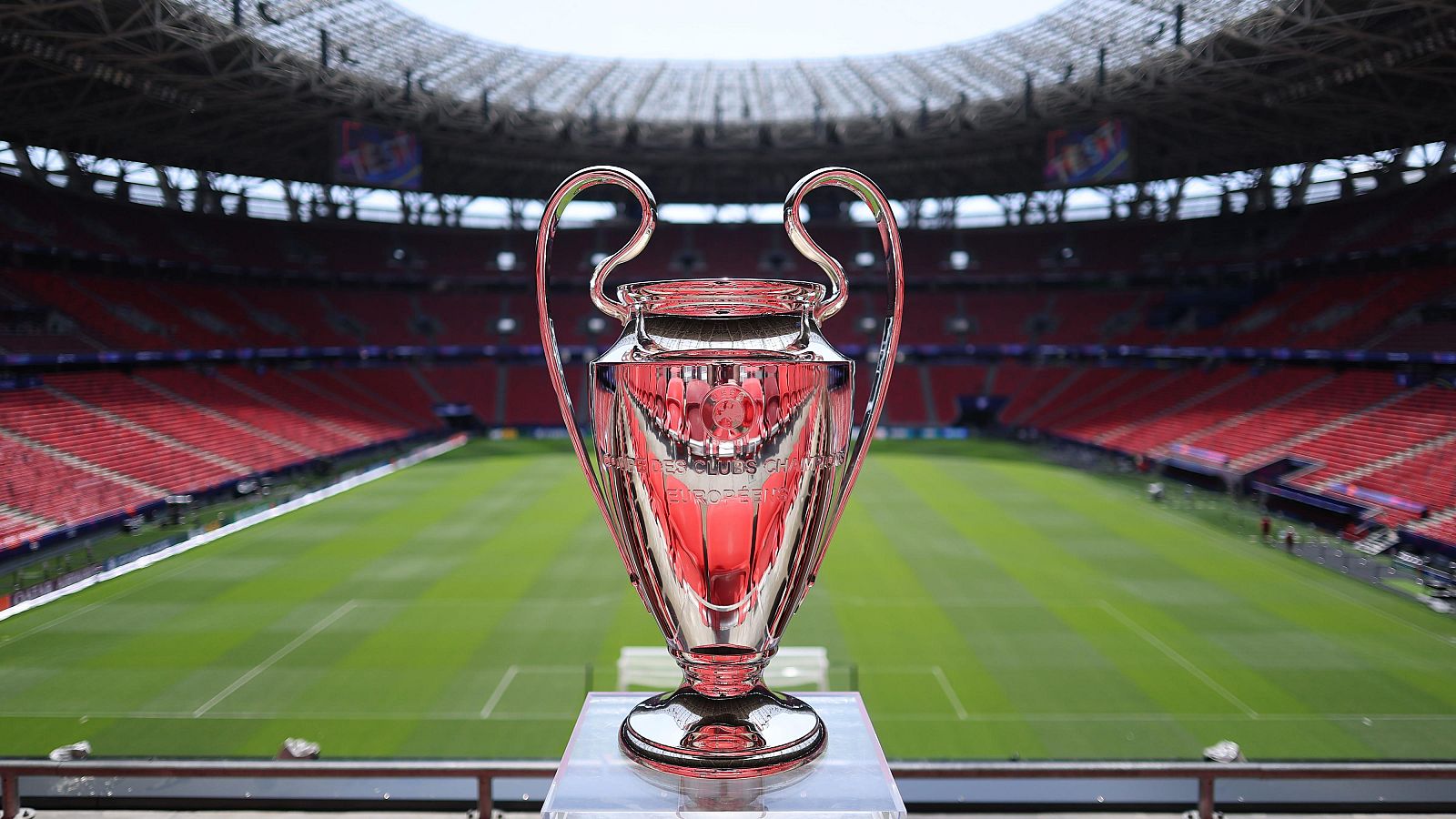 Trofeo de la UEFA Champions League plateado, con asas, sobre pedestal transparente en estadio vacío con asientos rojos. Grabados: "EUROPEAN CLUBS CHAMPION" y "COUPE DES CLUBS CHAMPIONS EUROPÉENS".