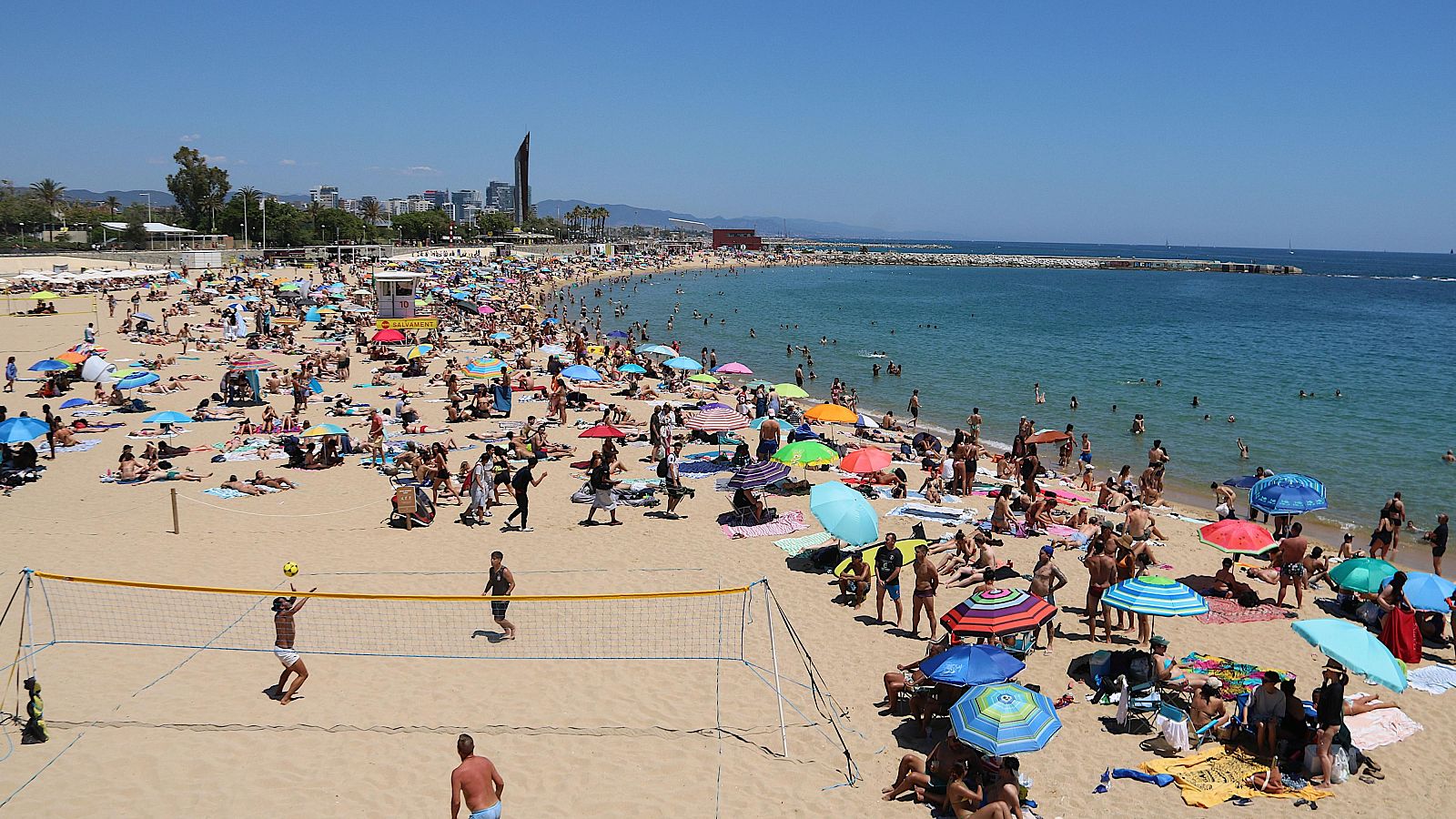 Playa urbana concurrida, voleibol en primer plano, multitud disfrutando, sombrillas, caseta de socorrista, espigón y rascacielos en el horizonte.