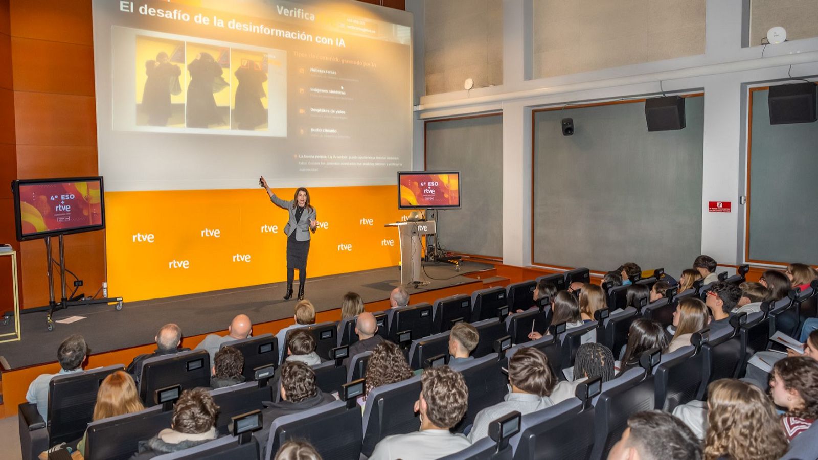 Montserrat Rigall da una masterclass sobre desinformación con IA a estudiantes de 4º ESO+RTVE en un auditorio con fondo naranja de RTVE. La pantalla principal muestra "El desafío de la desinformación con IA".