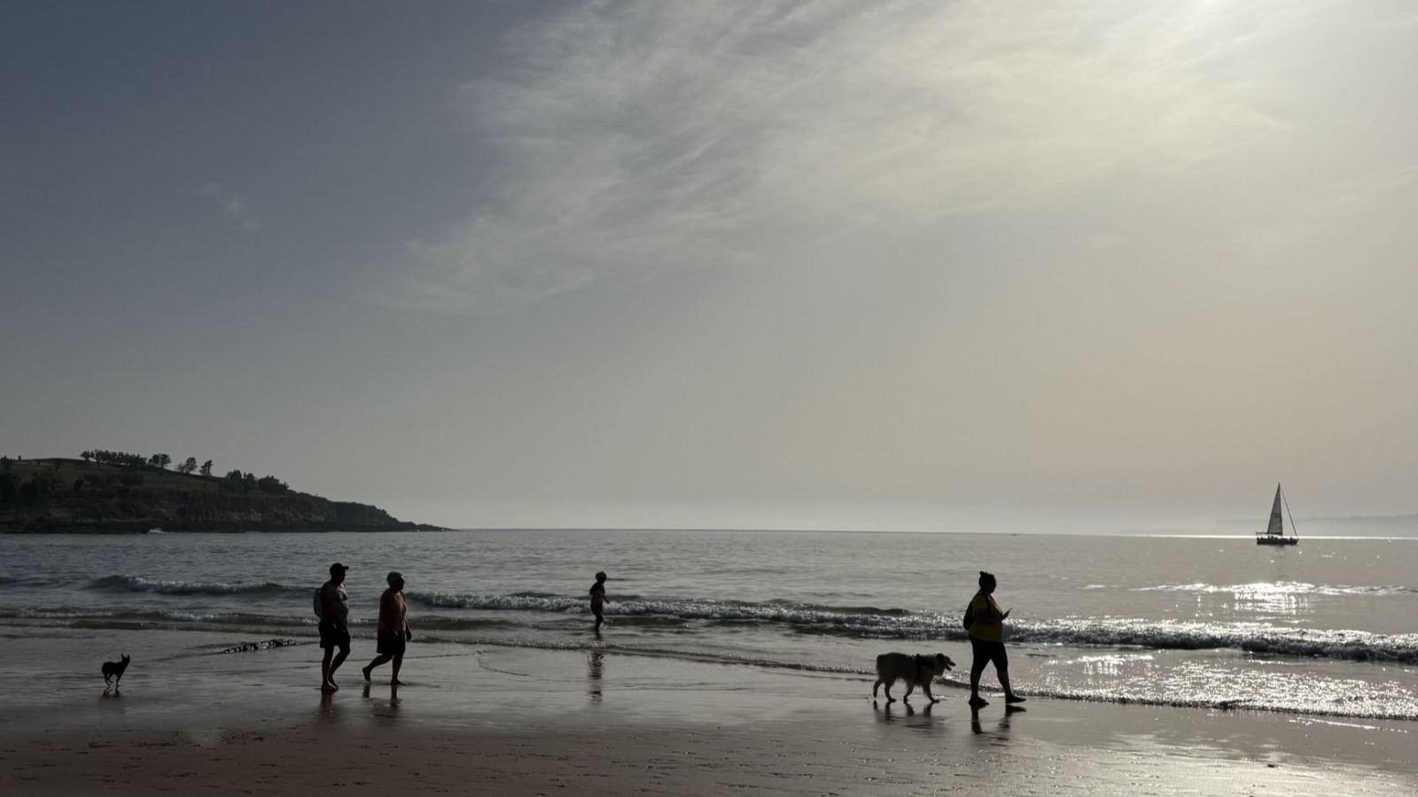 Siluetas de personas y perros pasean por la orilla de una playa al amanecer, con el sol bajo y un velero en el horizonte.