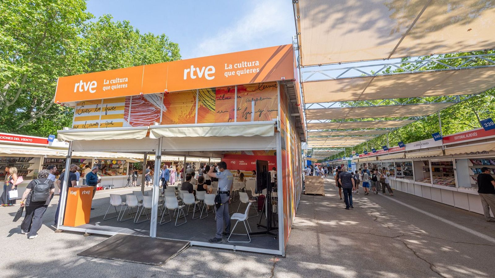 Vista panorámica de la Feria del Libro con un prominente stand de RTVE naranja y casetas de editoriales blancas llenas de libros. Multitud diversa pasea bajo toldos beige en una avenida arbolada y soleada.