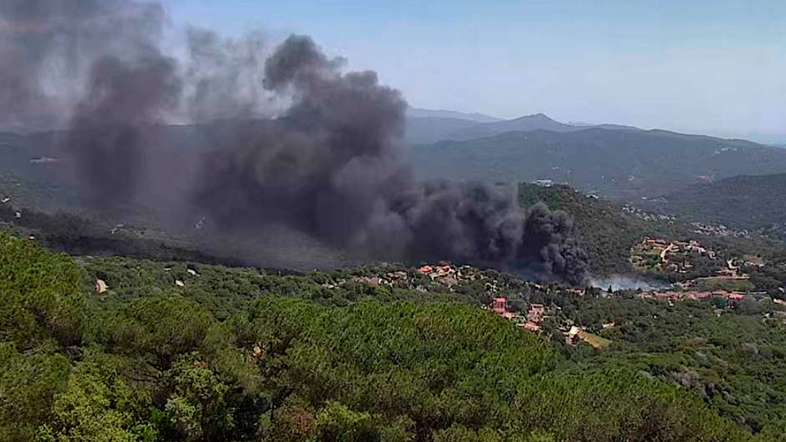 Columna de fum negre d'un incendi forestal que s'origina prop d'edificis en un paisatge muntanyós.