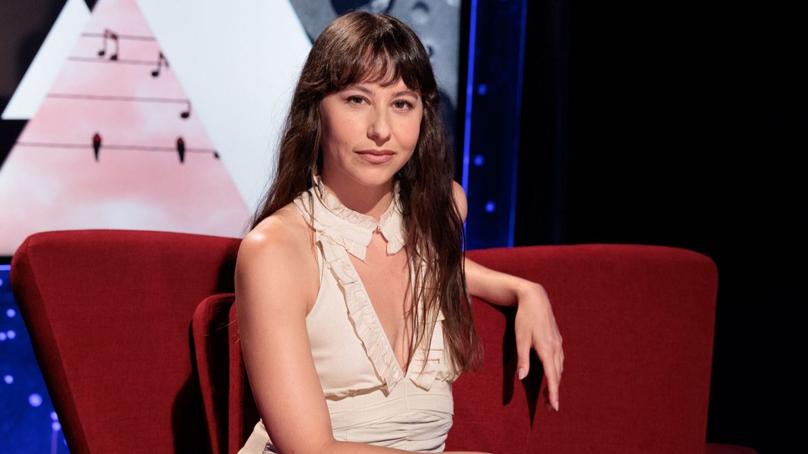 Irene Escolar, actriz española, sentada en sofá rojo, blusa de volantes, fondo de estudio con panel blanco y notas musicales.