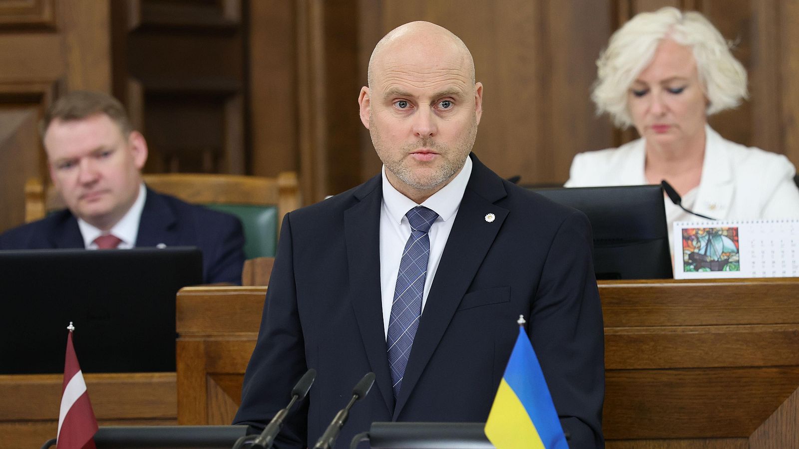 Primer ministro de Letonia, Andris Kulbergs, habla en el Parlamento. Banderas de Letonia y Ucrania presentes. Ambiente formal.