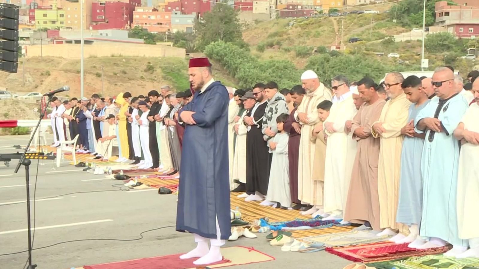 Oración colectiva en Ceuta con hombres y niños vestidos de forma tradicional, liderada por un imán. Se aprecian alfombras de oración, un equipo de sonido y un paisaje urbano al fondo.