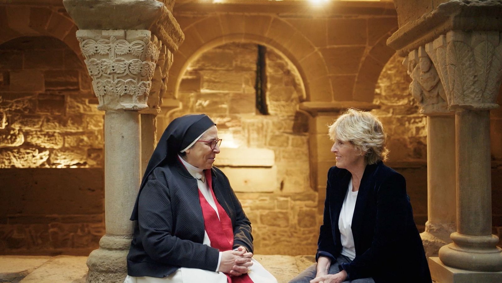 Sor Lucía Caram y Mercedes Milá conversan en un interior de piedra antigua, iluminado cálidamente. Sor Lucía lleva hábito y gafas; Mercedes, pelo rubio y americana azul.