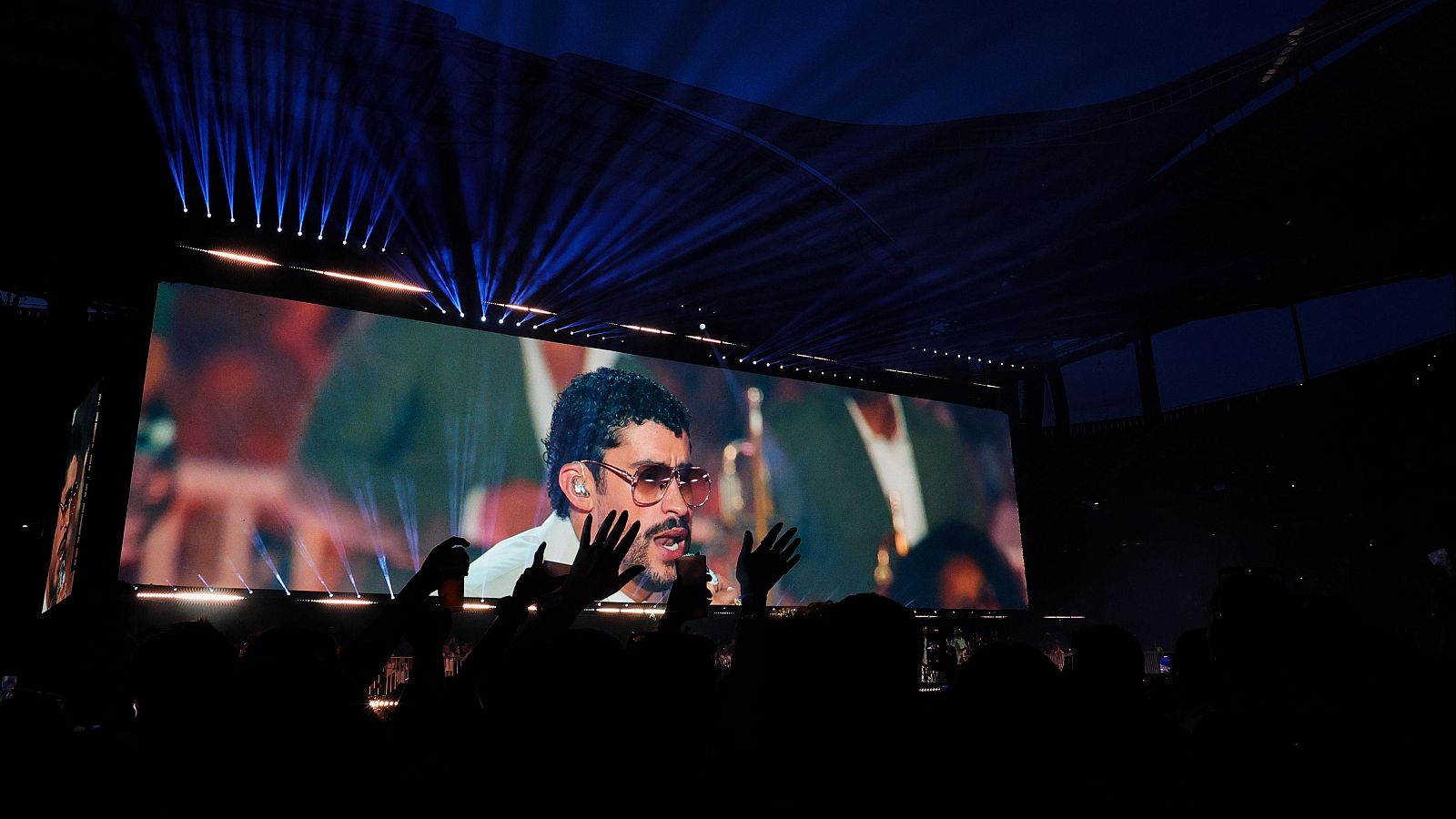 Concierto de Bad Bunny en Lisboa. El artista aparece en pantalla con gafas de sol, rodeado de una multitud eufórica y luces azules.