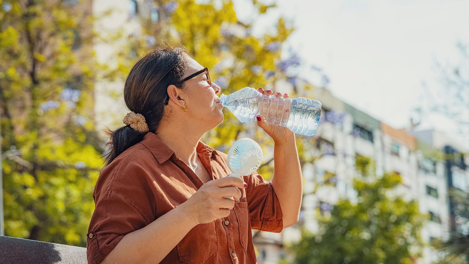 Una mujer bebe agua y se refresca con un ventilador portátil