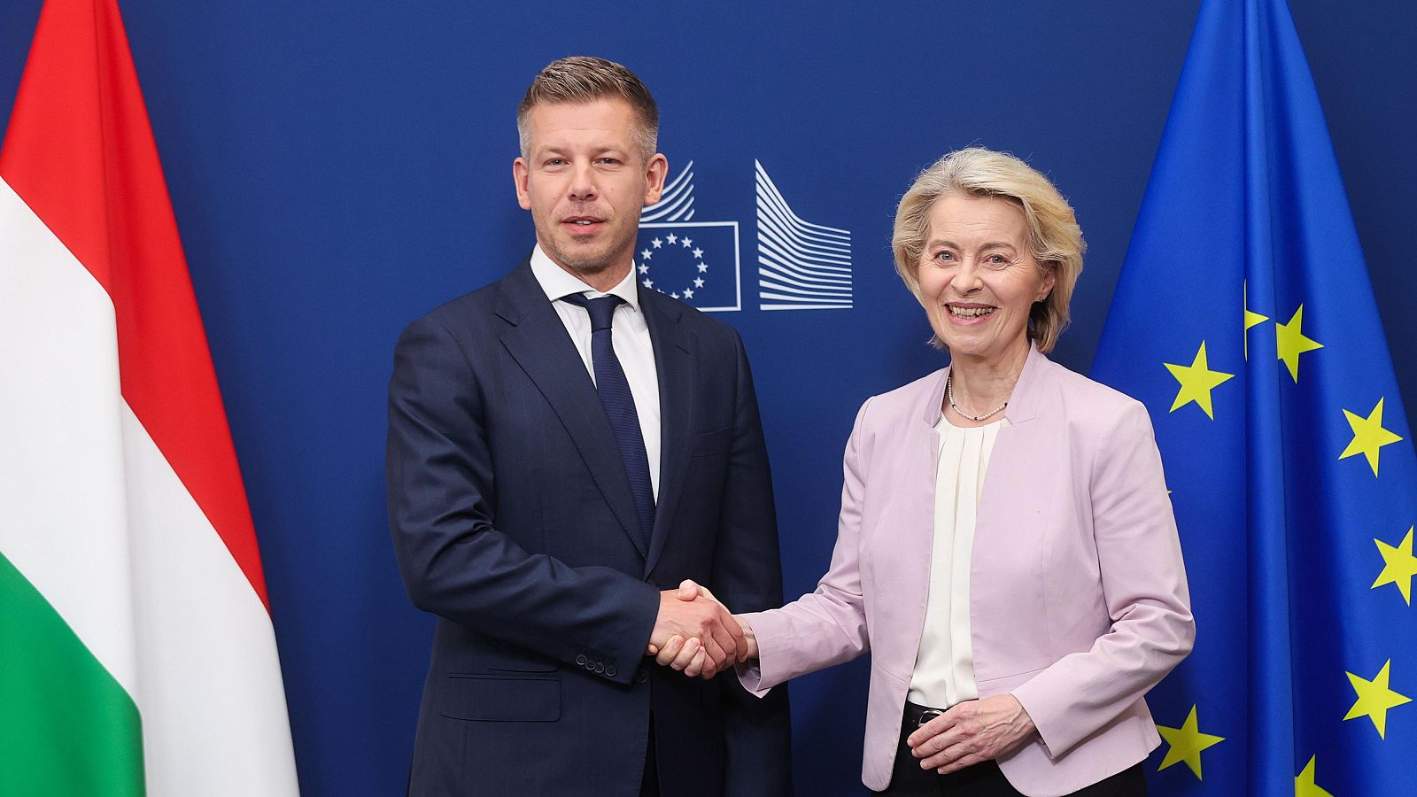 Peter Magyar y Ursula von der Leyen se estrechan la mano en un encuentro diplomático, con las banderas de Hungría y la UE de fondo.
