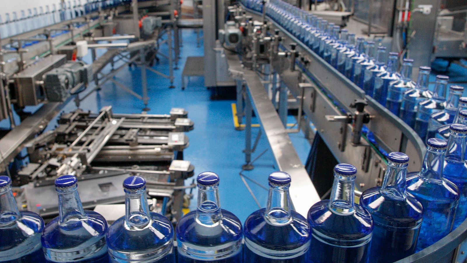 Planta embotelladora de Fuensanta en Asturias, con botellas azules en cintas transportadoras y maquinaria gris sobre suelo azul brillante. Proceso automatizado.