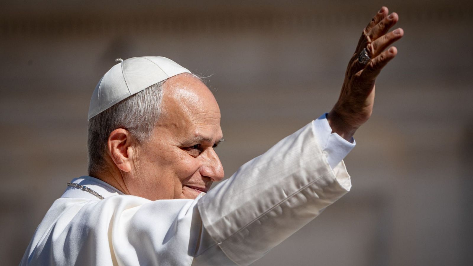 Primer plano del Papa Francisco de perfil, con sotana blanca, mano levantada en gesto de saludo y anillo en el dedo.