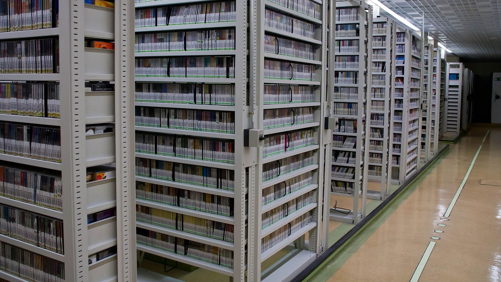 Pasillo de archivo con estanterías móviles de alta densidad, cajas blancas con etiquetas verdes y suelo beige. Iluminación fluorescente en el techo.
