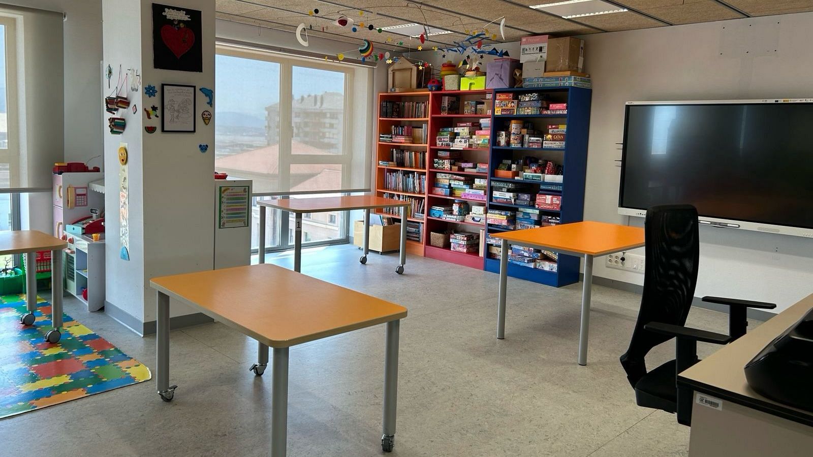 Aula hospitalaria con mobiliario colorido, estanterías de juegos y libros, y pantalla interactiva, iluminada por luz natural.