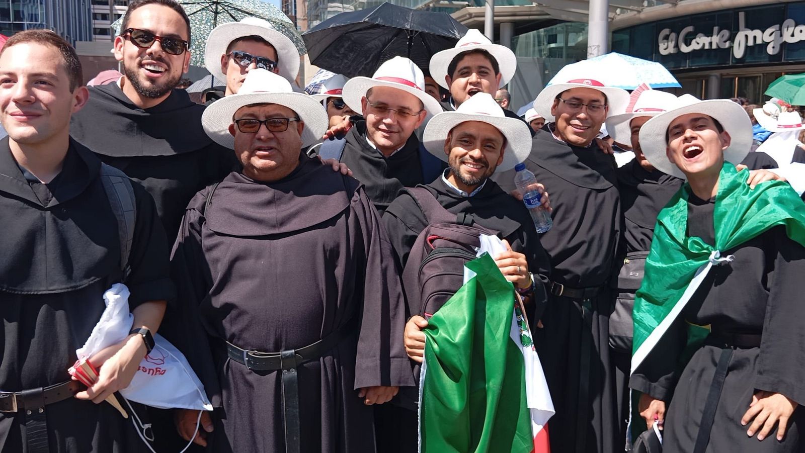 Jóvenes agustinos recoletos con hábitos y sombreros blancos, algunos portando la bandera de México, esperan en un día soleado frente a un edificio con el logo de "El Corte Inglés".