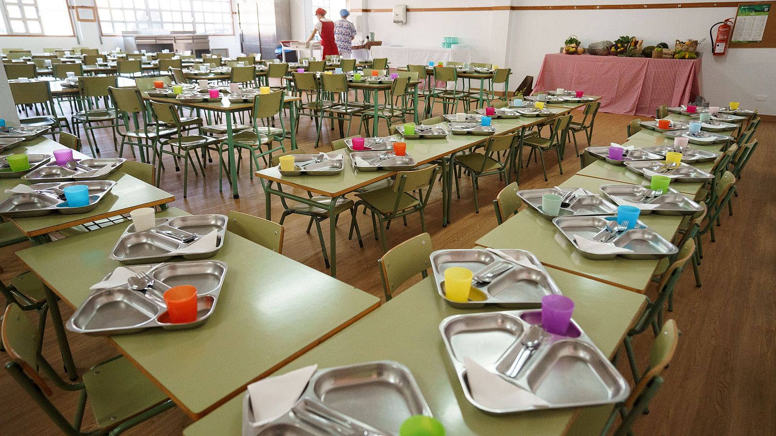 Comedor escolar preparado con mesas, bandejas, cubiertos y vasos de colores. Frutas y verduras frescas en una mesa. Personal uniformado en la zona de servicio. Ambiente luminoso y ordenado.