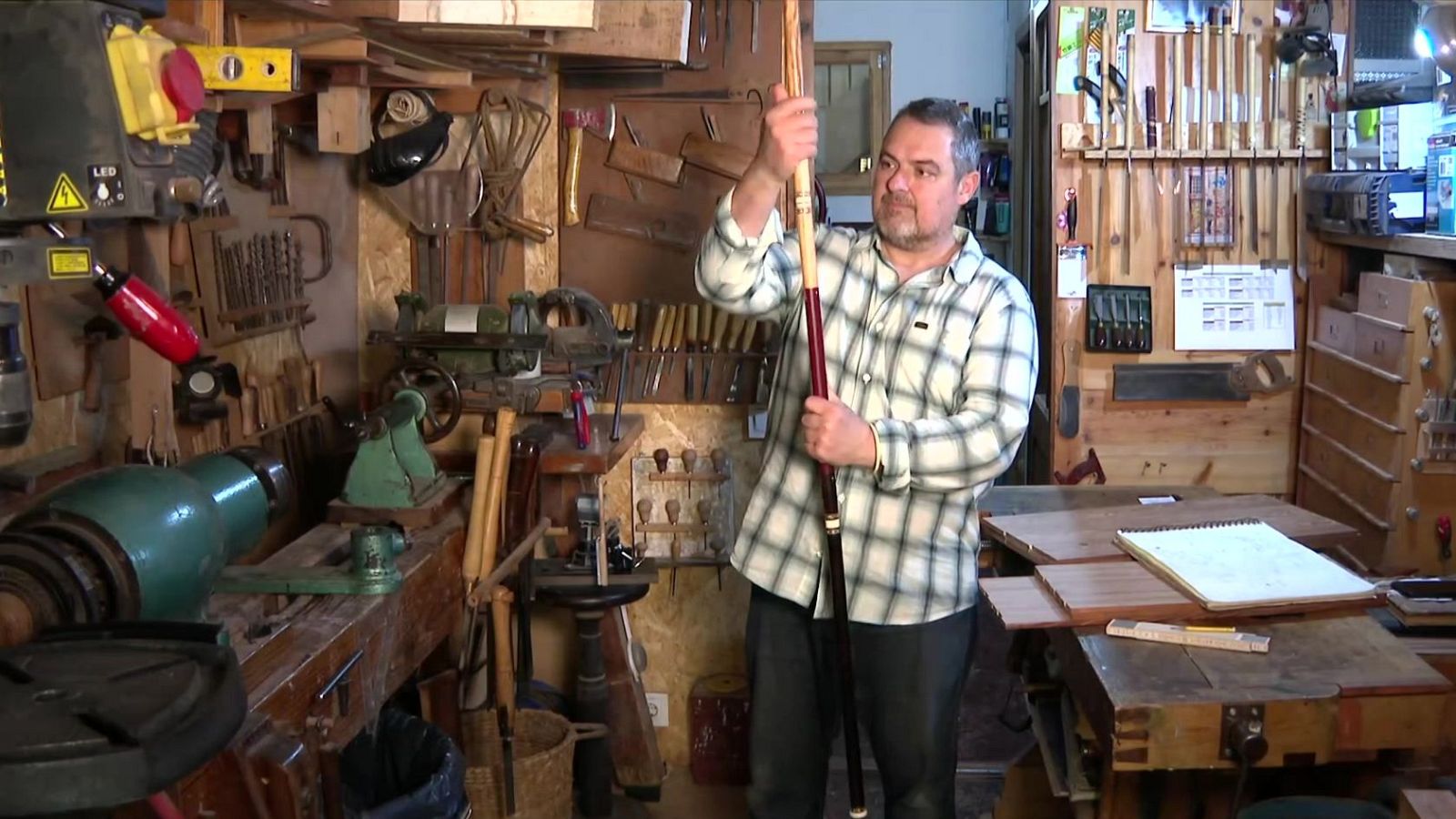 Òscar Ollé, ebanista, posa en el seu taller amb un bàcul de fusta tallat per ell mateix. L'espai està ple d'eines i maquinària d'ebenisteria.