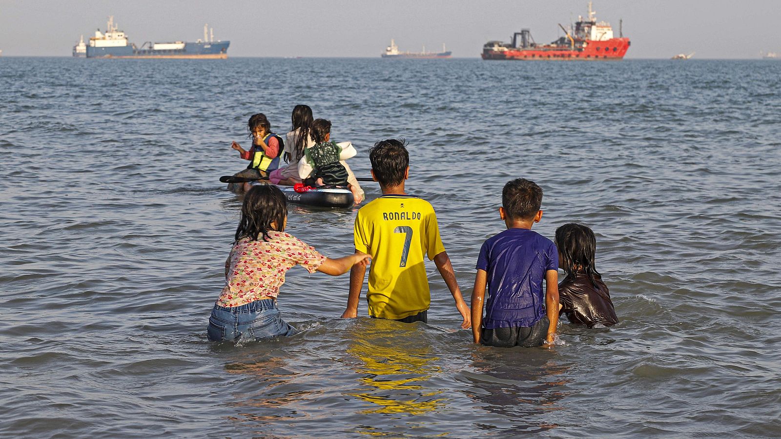 Niños iraníes juegan en la playa de Suru, Bandar Abbas. Uno lleva una camiseta de "RONALDO 7". Embarcaciones navegan en el horizonte azul verdoso.