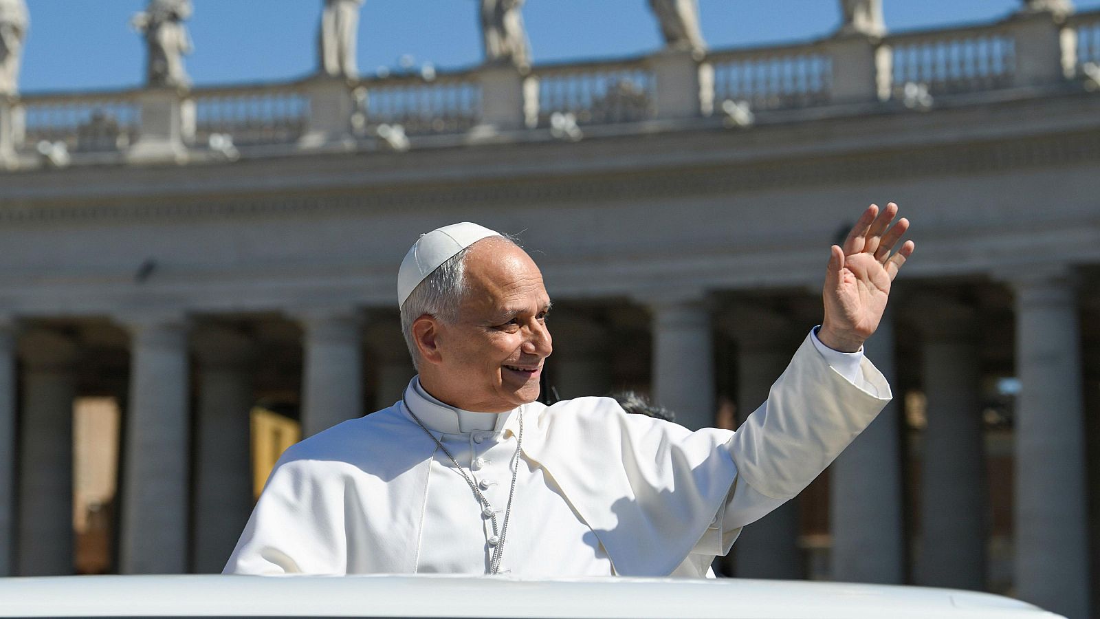 El Papa León XIV saluda sonriente desde un vehículo, con la Plaza de San Pedro de fondo. Su vestimenta papal y la arquitectura clásica son visibles.