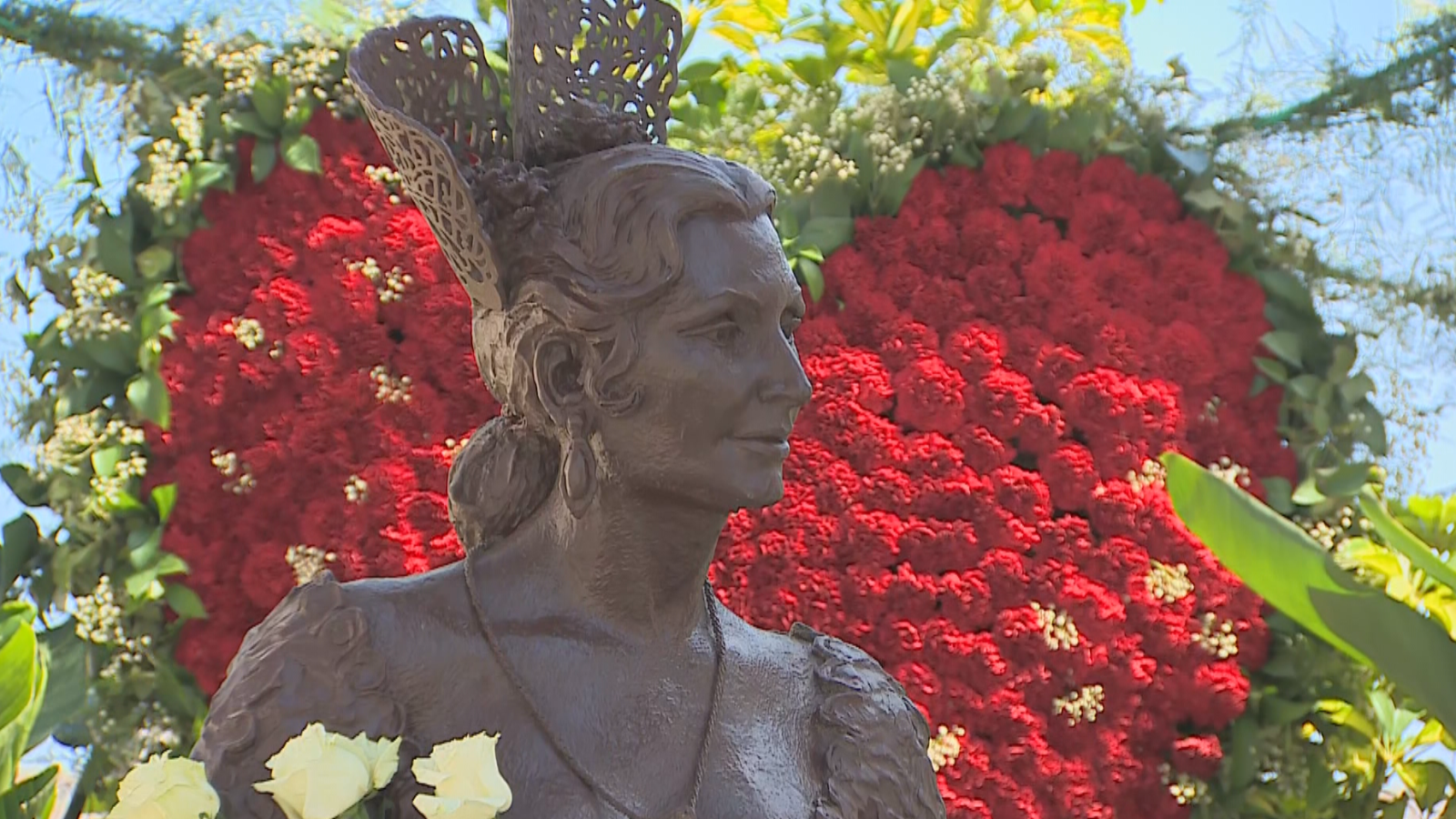 Busto de bronce de Rocío Jurado con tocado, sobre un corazón de flores rojas y blancas. Cielo azul al fondo.