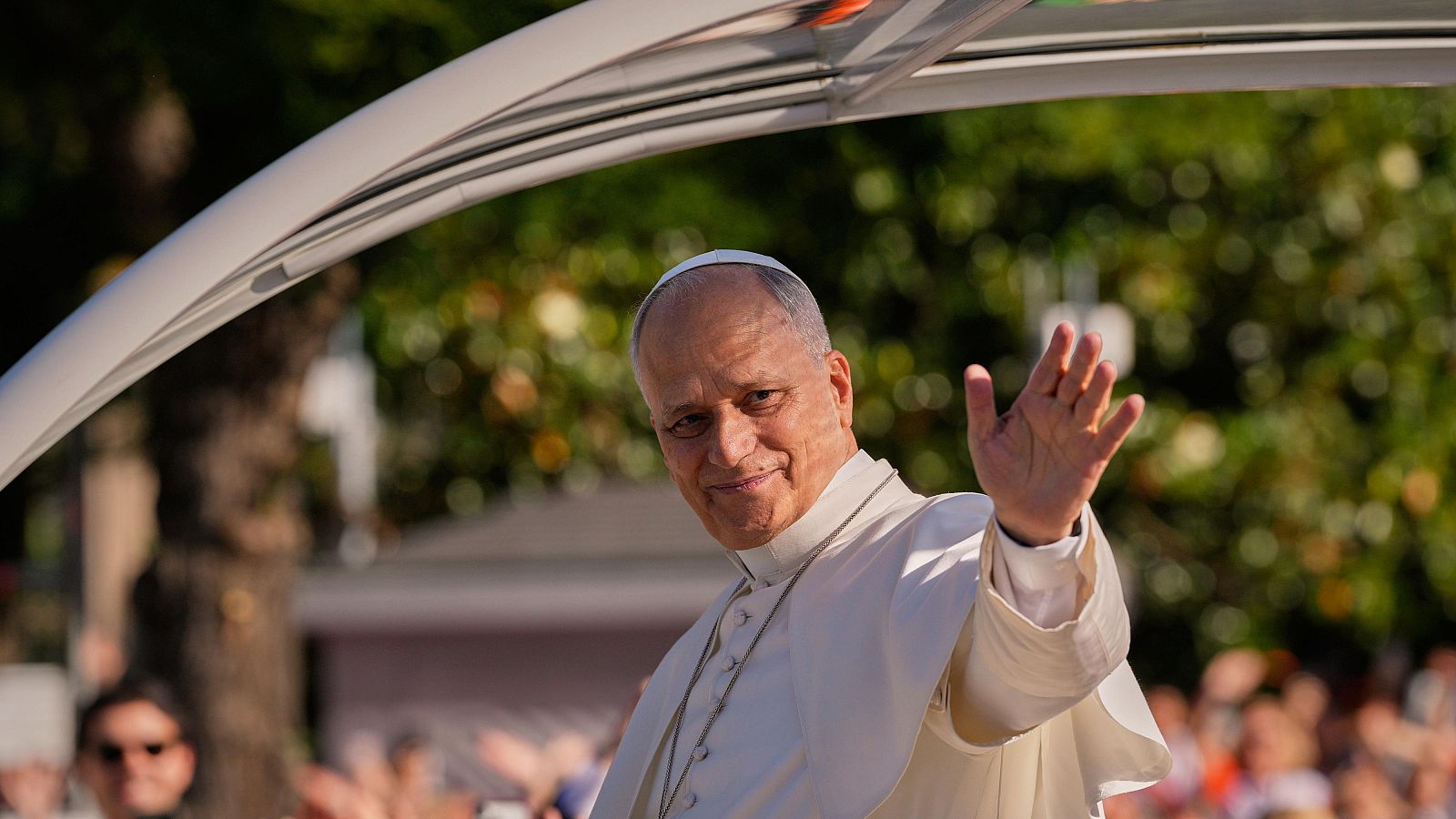 El Papa León XIV saluda sonriente desde un papamóvil, con solideo blanco y cruz pectoral, ante una multitud y vegetación.