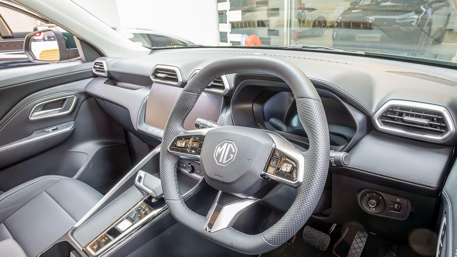 Vista del interior de un coche moderno: volante multifunción con logo MG, salpicadero con dos pantallas digitales, consola central y panel de puerta. Destacan detalles en plateado y texturas de cuero.