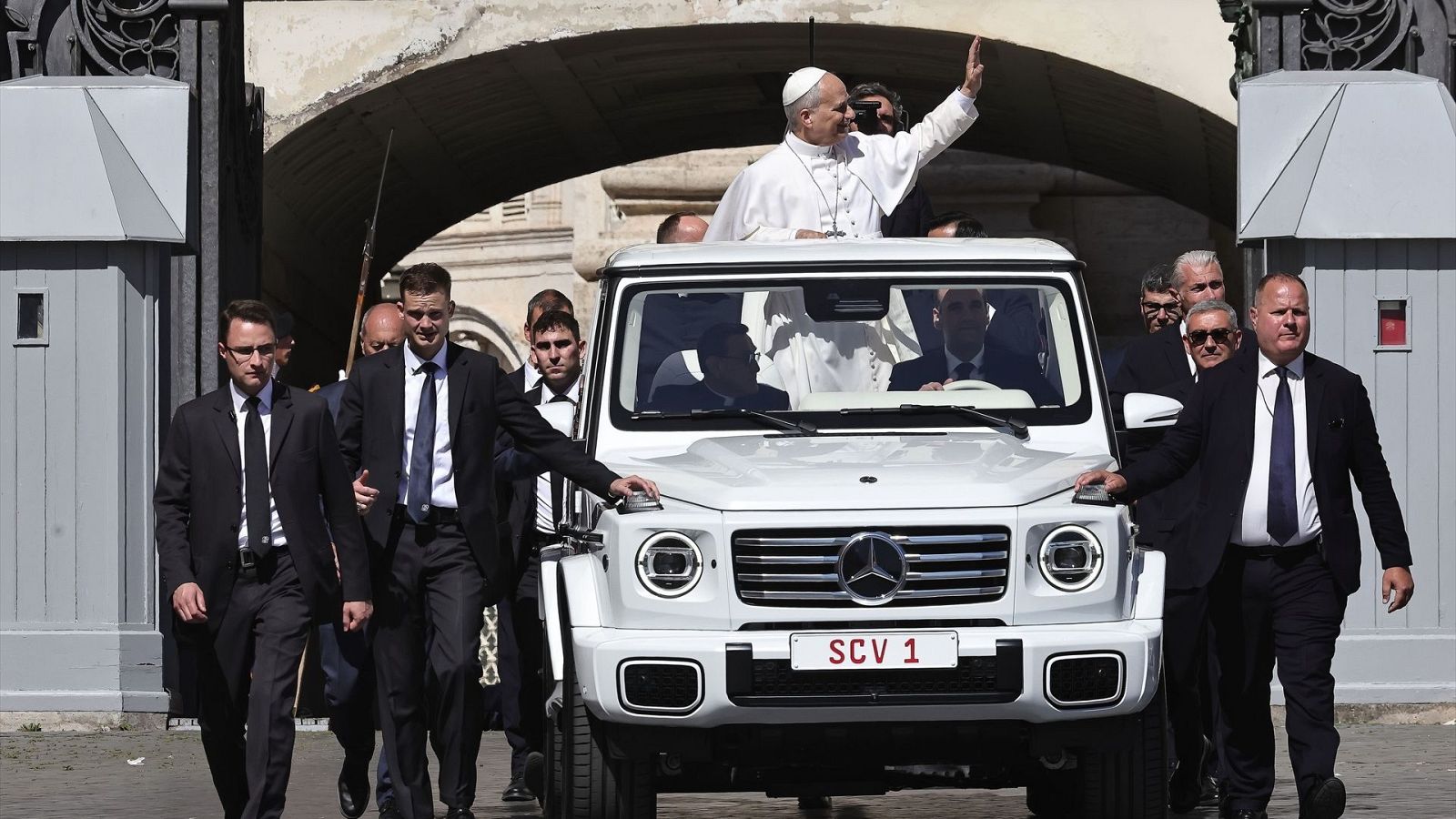 El Papa Francisco saluda desde su papamóvil blanco "SCV 1" al emerger de un arco de piedra en la Plaza de San Pedro, escoltado por seguridad.