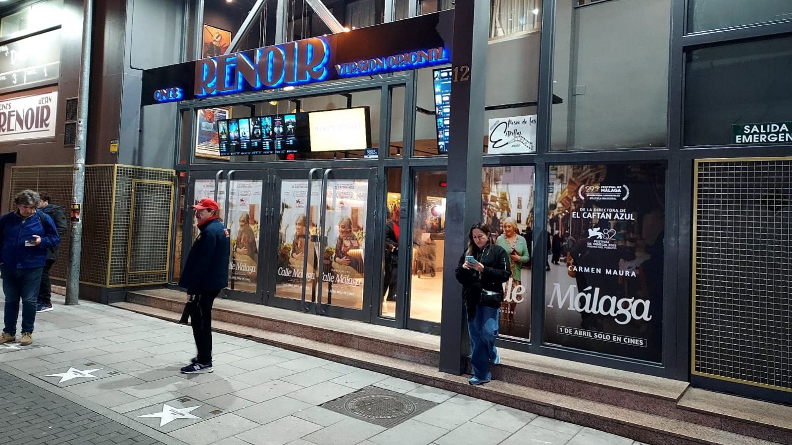 Fachada nocturna de los Cines Renoir con letreros de neón, personas en la acera y entrada, y un póster de la película "Málaga" con Carmen Maura.