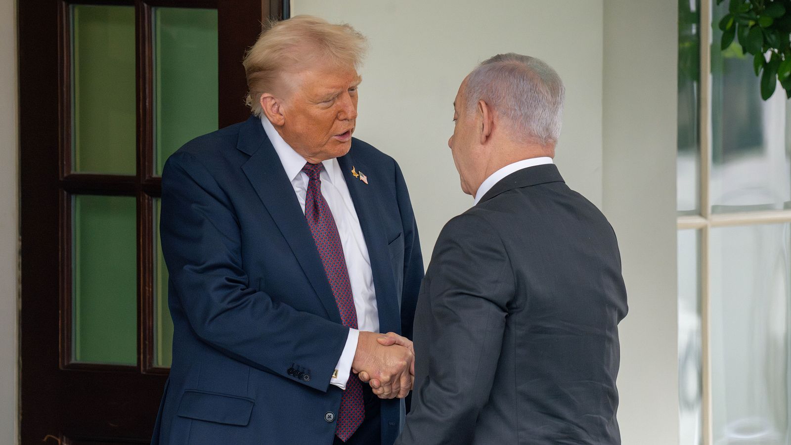 Donald Trump y Benjamin Netanyahu se dan la mano en un encuentro. Trump viste traje azul y corbata granate; Netanyahu, traje oscuro. El fondo es luminoso con vegetación.