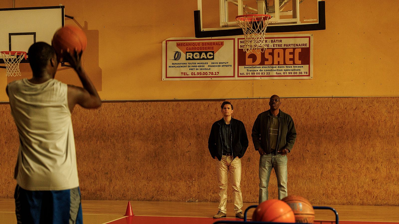 Tres hombres en una cancha de baloncesto interior. Uno lanza, dos observan. Carteles en francés anuncian servicios de mecánica y construcción. Fotograma de 'El sueño americano'.