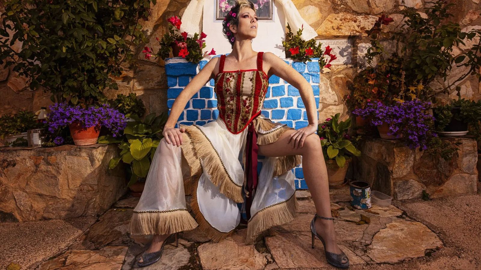Karmento, cantante española, sentada en pose artística con corsé rojo y beige, falda blanca con flecos, tacones oscuros, cabello rubio platino con flores rosas, en un patio rústico con pared de piedra y vegetación.