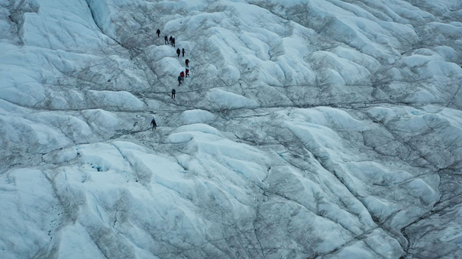 Expedición en un glaciar remoto, con figuras humanas avanzando sobre hielo accidentado y grietas oscuras. Vista aérea que resalta la majestuosidad y escala del paisaje glacial.