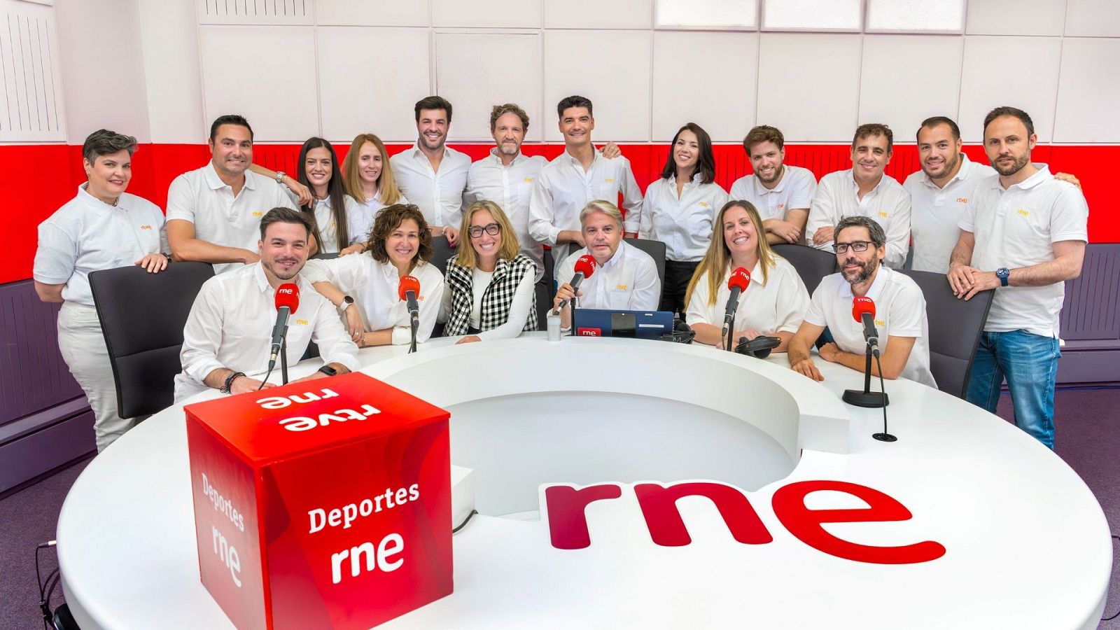 Equipo de RNE Deportes posa en estudio de radio con mesa en forma de herradura y cubo con logo "Deportes rne". Siete sentados con micrófonos, diez de pie detrás.
