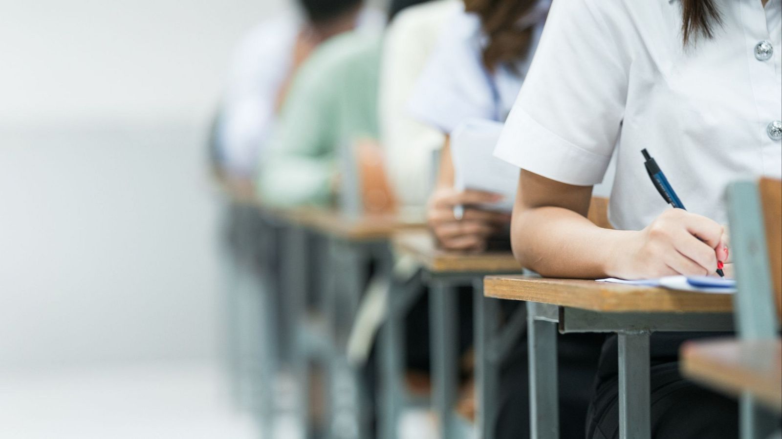 Primer plano de una mano con camisa blanca escribiendo con bolígrafo azul sobre papel en un examen, con otras personas concentradas al fondo.