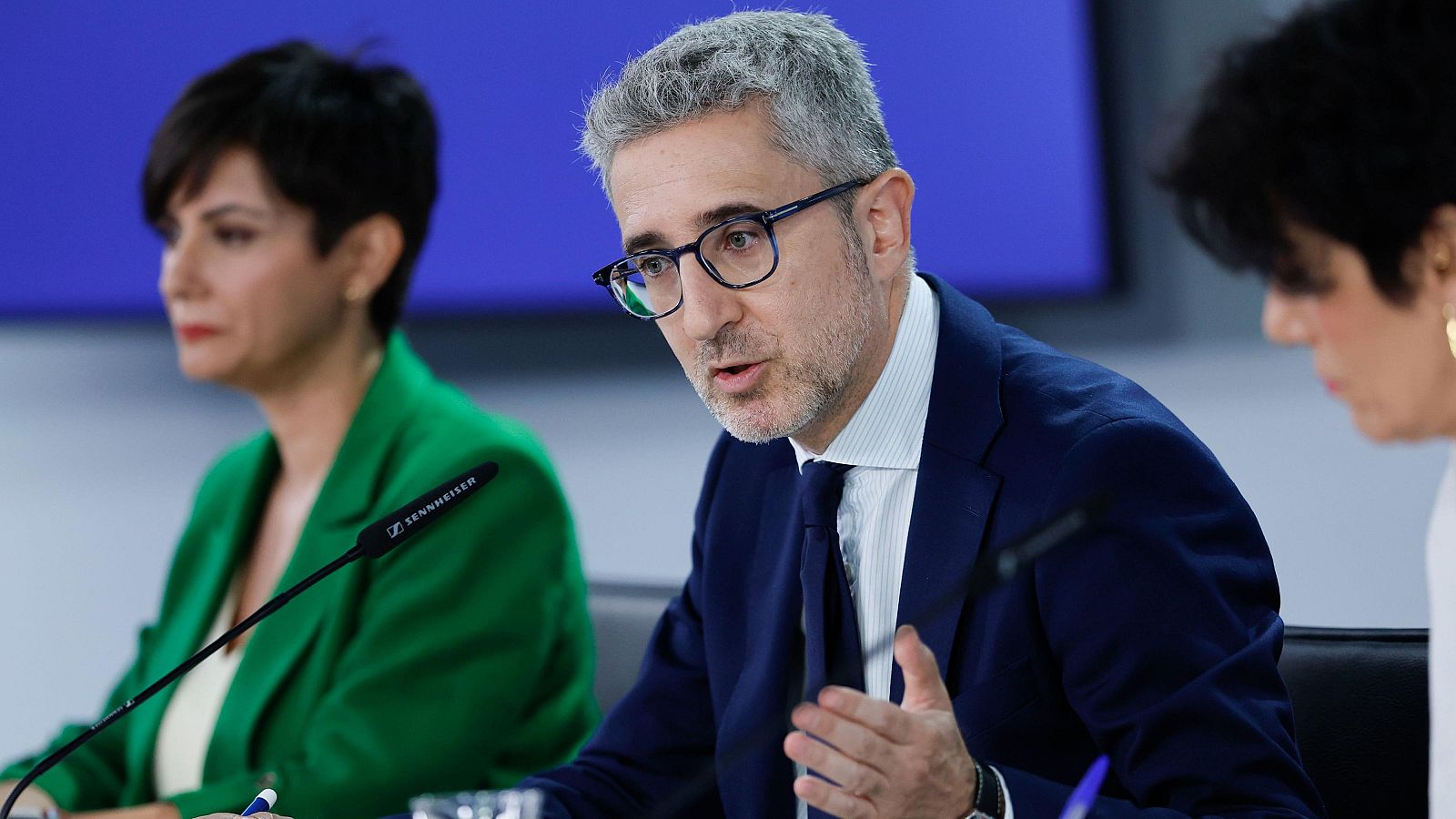 Rueda de prensa del Consejo de Ministros con Arcadi España, Elma Saiz e Isabel Rodríguez. Se observan micrófonos Sennheiser, vasos de agua y bolígrafos sobre la mesa.