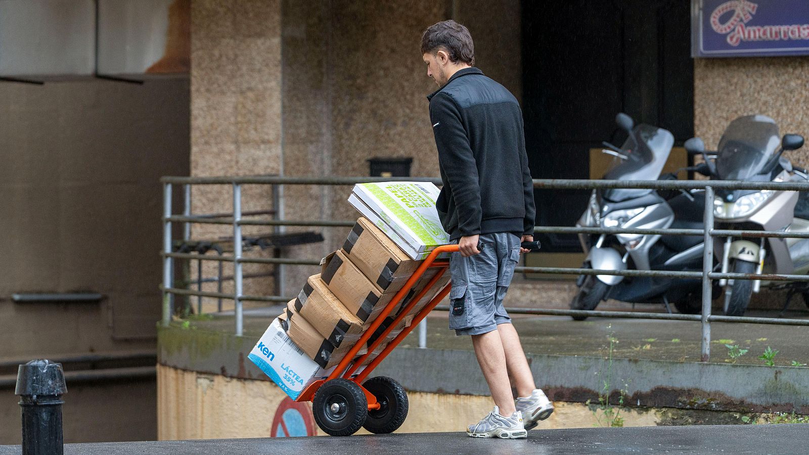 Hombre empuja carretilla naranja con cajas y paquete blanco/verde sobre suelo mojado. Fondo: barandilla, edificio con cartel "Amarras" y dos motos.
