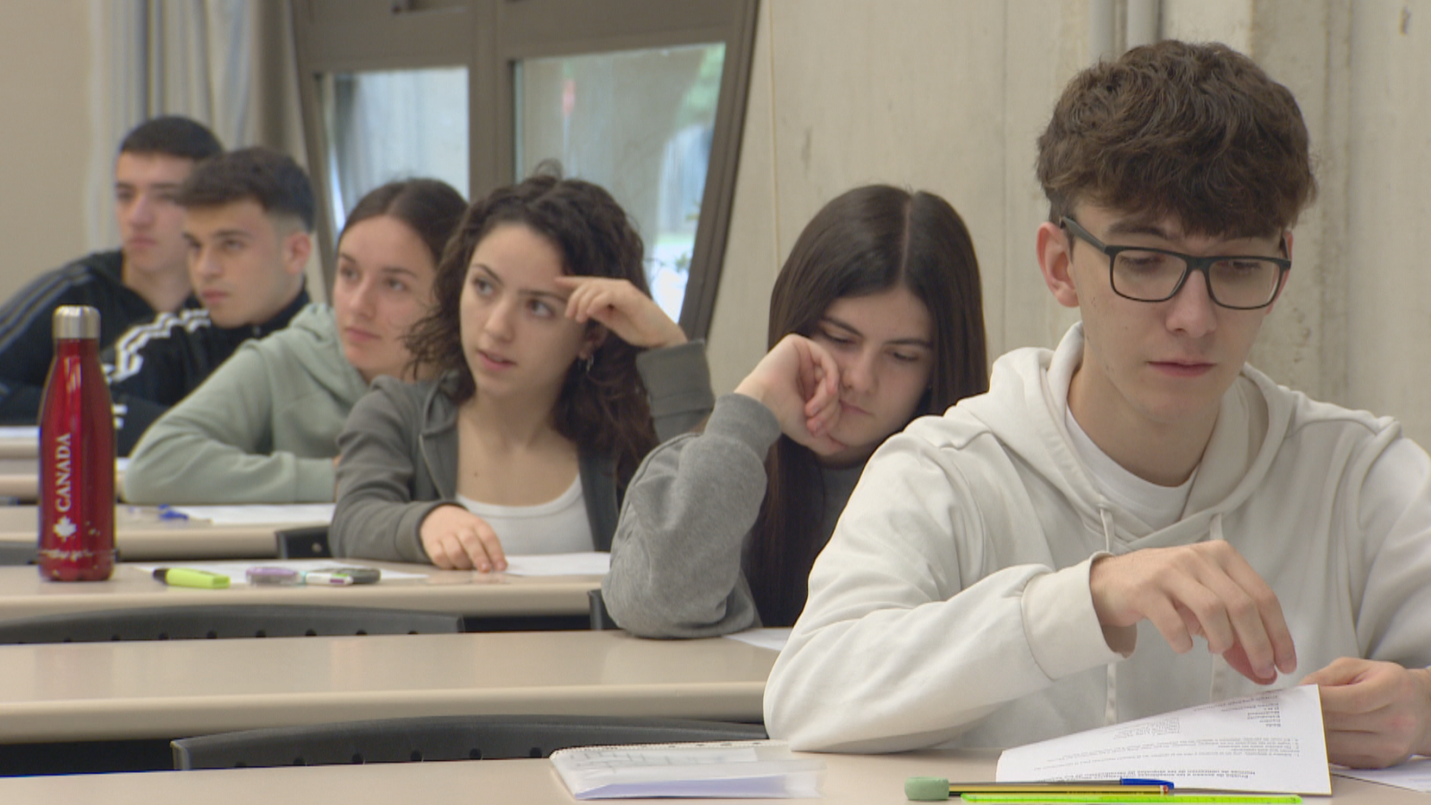 Estudiantes concentrados en un aula, con materiales de estudio sobre sus pupitres, momentos antes de un examen. Se aprecian detalles de vestimenta y expresiones.