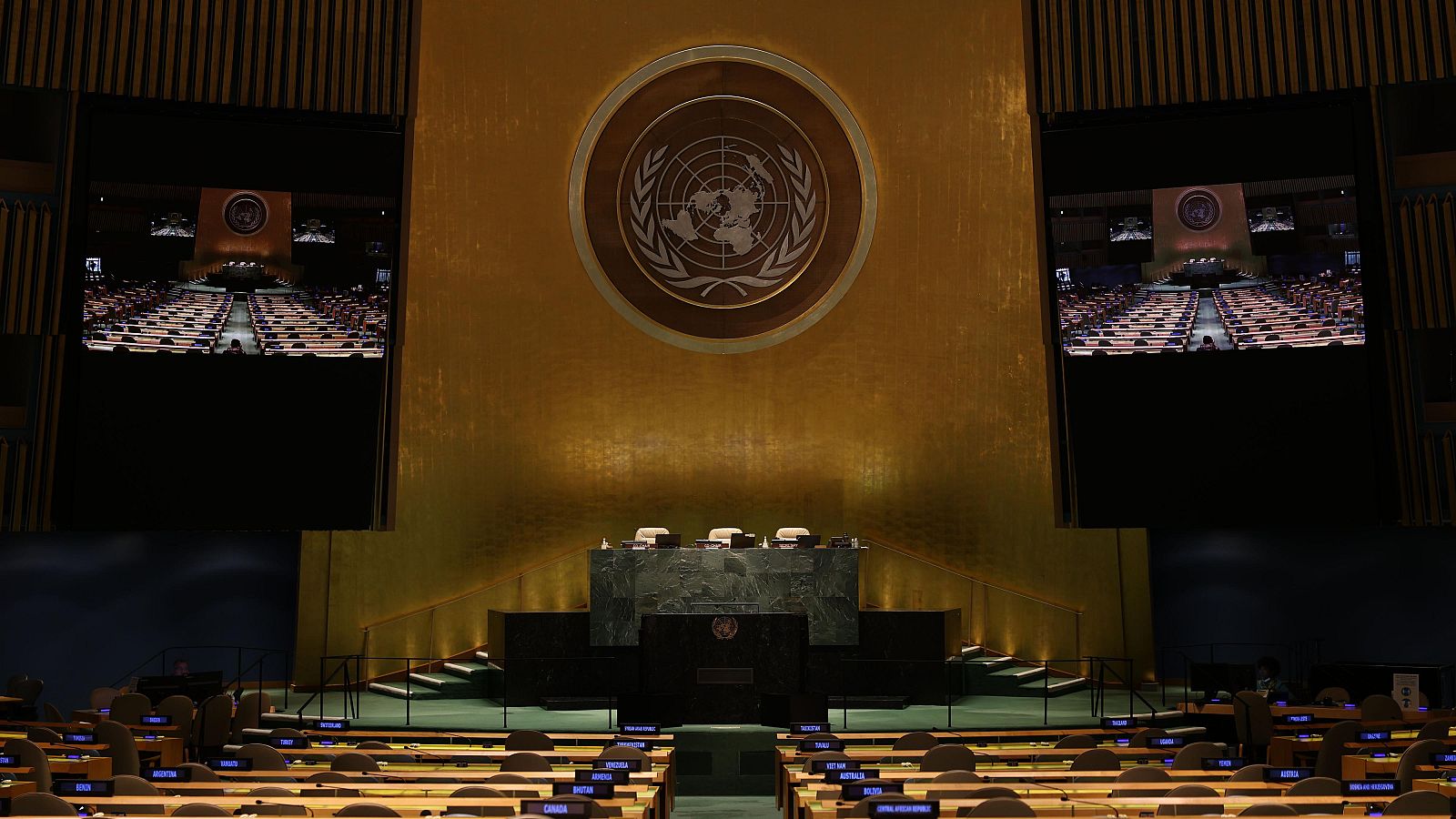 Interior de la Cámara de la Asamblea General de la ONU, vacío, con el emblema de la ONU al fondo, escritorios y sillas en semicírculo, y pantallas mostrando la sala.