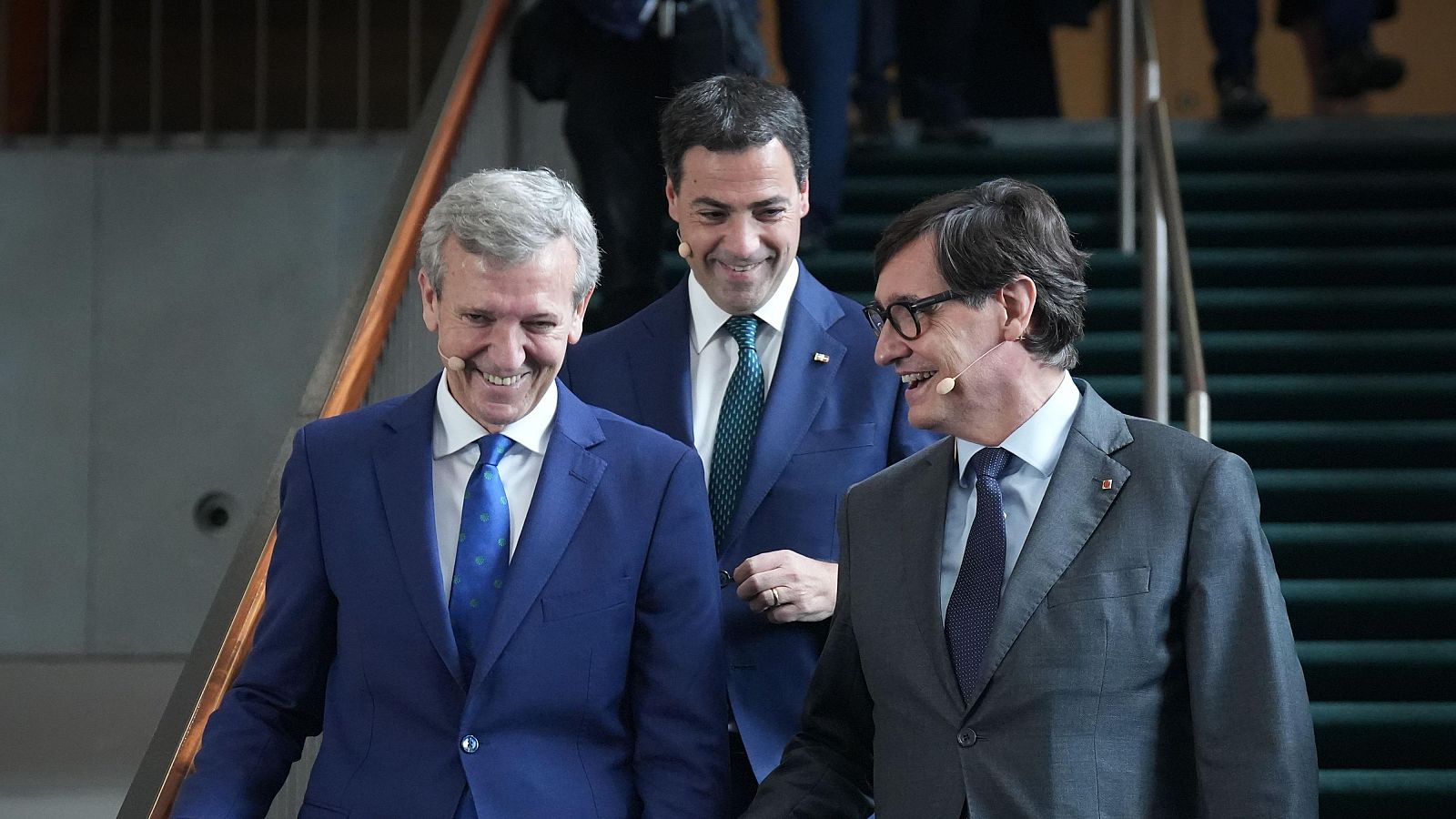 Alfonso Rueda, Imanol Pradales y Salvador Illa, políticos españoles, conversan sonrientes mientras descienden unas escaleras con trajes formales.