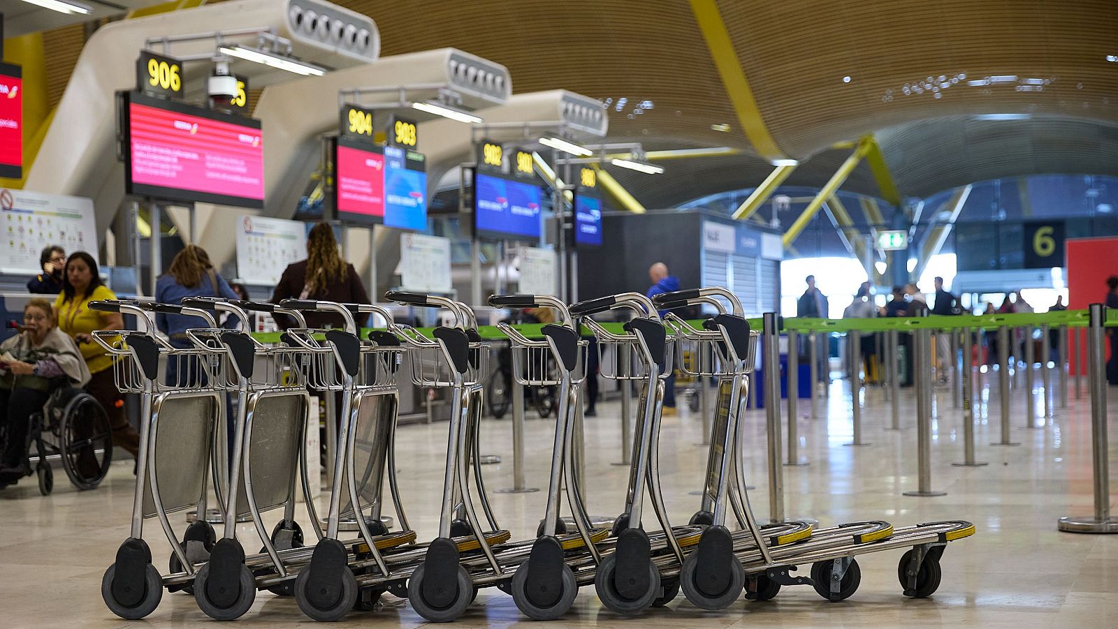 Terminal de aeropuerto con carros portaequipajes, mostradores de facturación con pantallas de Iberia y British Airways, y personas moviéndose por el espacio.