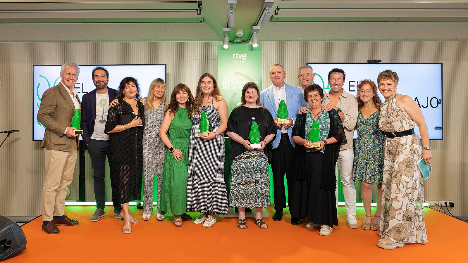 Doce personas celebran los V Premios El Escarabajo Verde de RTVE en un escenario con suelo naranja y fondo verde. Varios asistentes, como Jacobo Bergareche y Elisenda Roca, sostienen trofeos verdes.