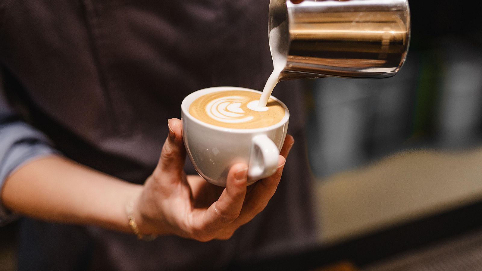 Preparación de café con leche con latte art. Una mano vierte leche espumosa sobre un café en una taza, mientras otra mano sostiene la taza. Se observa una pulsera dorada.