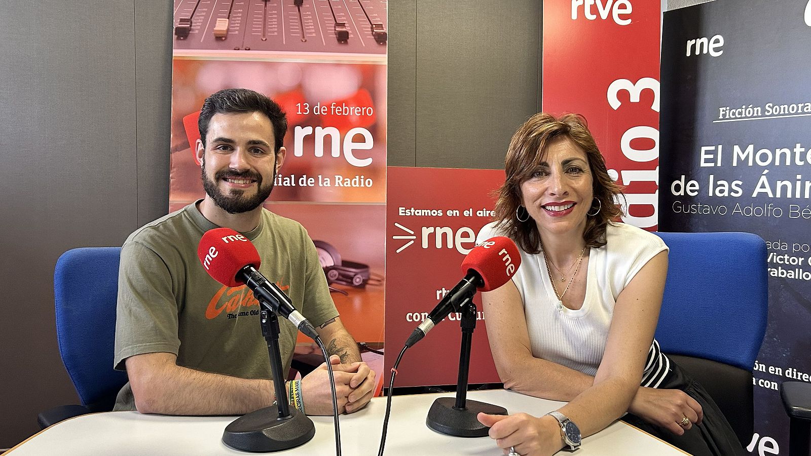 Carlos Huelin y Mamen Asencio, periodistas de RNE, posan en un estudio de radio con micrófonos y paneles informativos, incluyendo el "Día Mundial de la Radio".