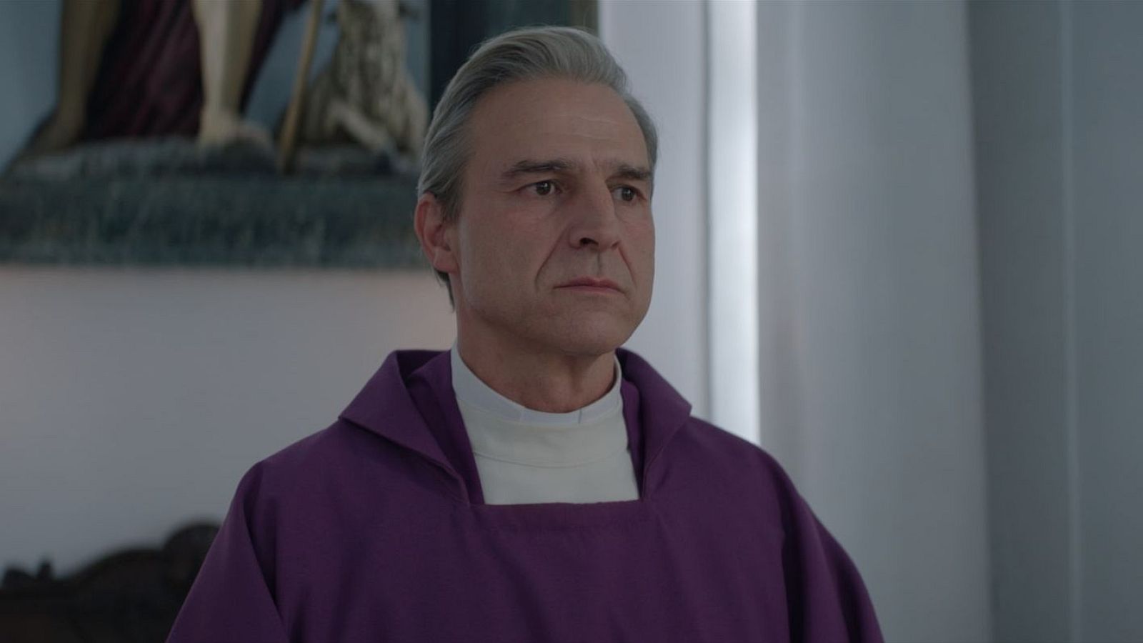 Karra Elejalde, con sotana morada y expresión seria, en un fotograma de 'La luz'. Se aprecian detalles de su cabello canoso y un fondo con elementos religiosos.