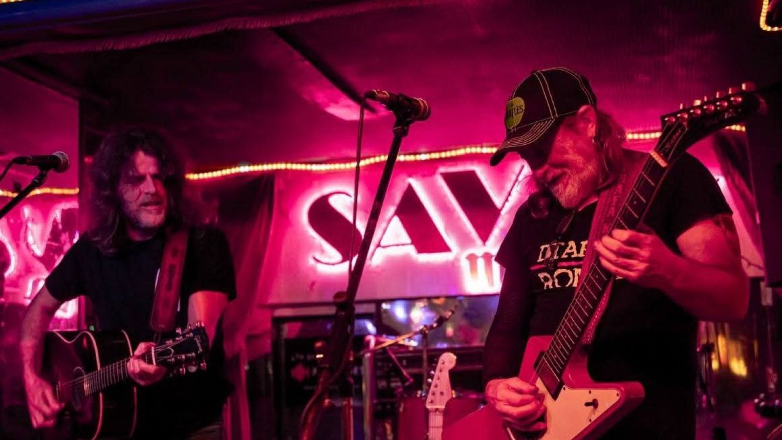 Dos músicos con barba y cabello largo tocan guitarras en un escenario con neones rosas y magenta. Un letrero de neón al fondo dice "SAV".