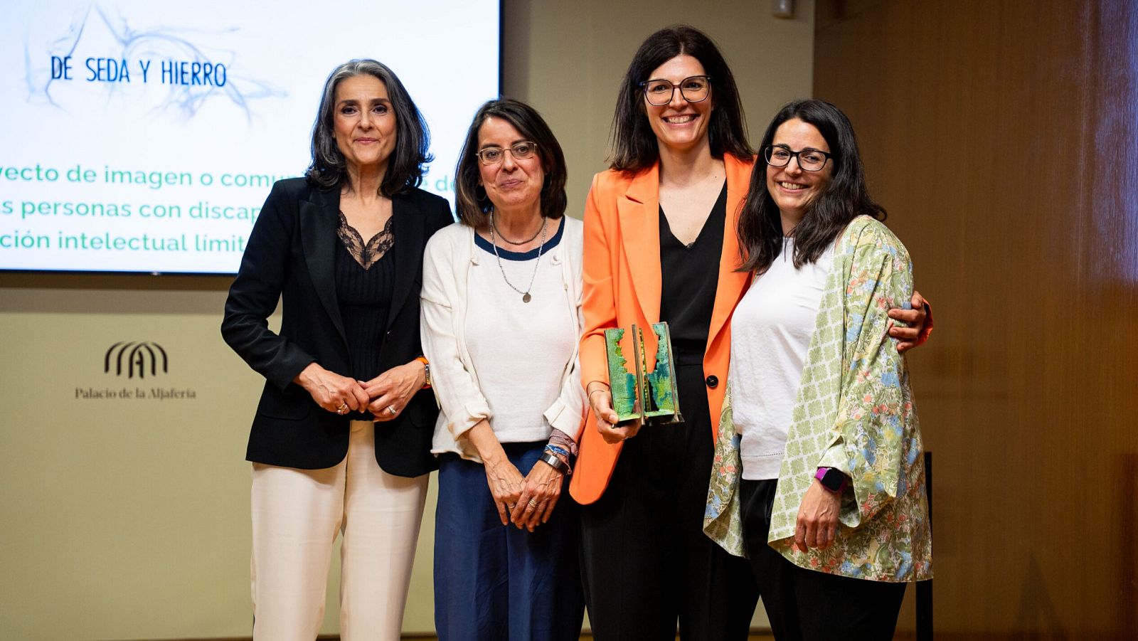 Cuatro mujeres sonríen en un evento. Patricia Martínez Marcos de León, directora, sostiene un premio. Cristina López Sacristán, realizadora, la abraza. Detrás, pantalla con "DE SEDA Y HIERRO".
